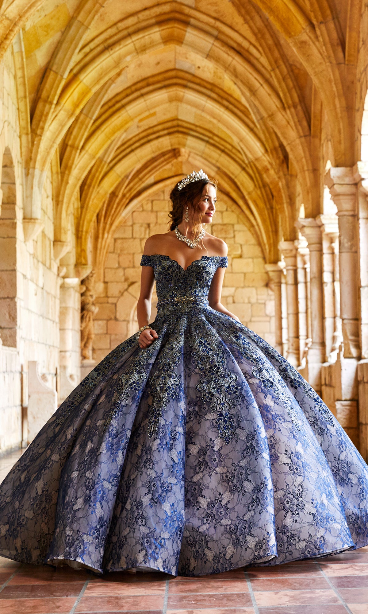 A woman in the Princesa Lace Ball Gown Quinceañera Dress PR11921 and a tiara stands in a stone archway with vaulted ceilings, gazing to the side.